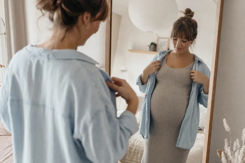 Gravid person provar en öppen skjorta över en mjuk klänning framför en spegel.