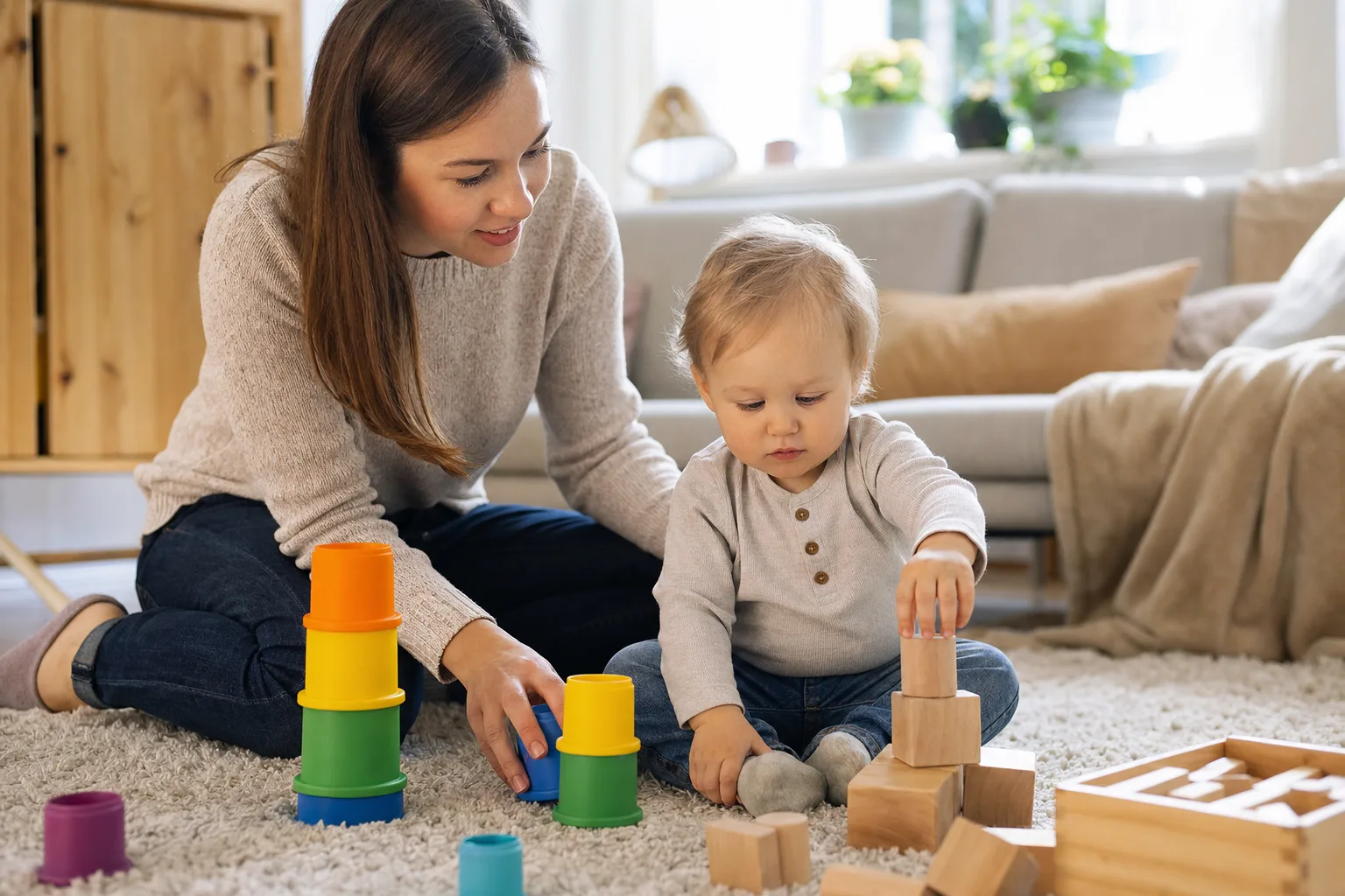 En förälder sitter på golvet med en 1-åring som leker med stapelmuggar och klossar.